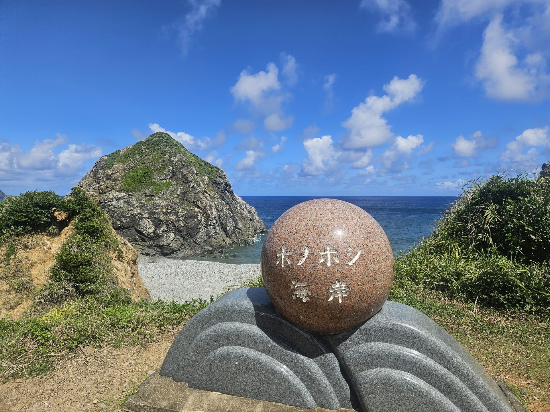 「ホノホシ海岸」と刻まれた球形の石碑と、背後の岩山、玉石の浜
