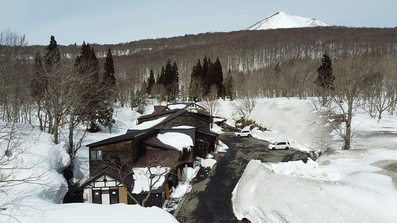 雪深い八甲田の山中に建つ谷地温泉の木造の宿を上空から捉えた空撮、奥に高田大岳