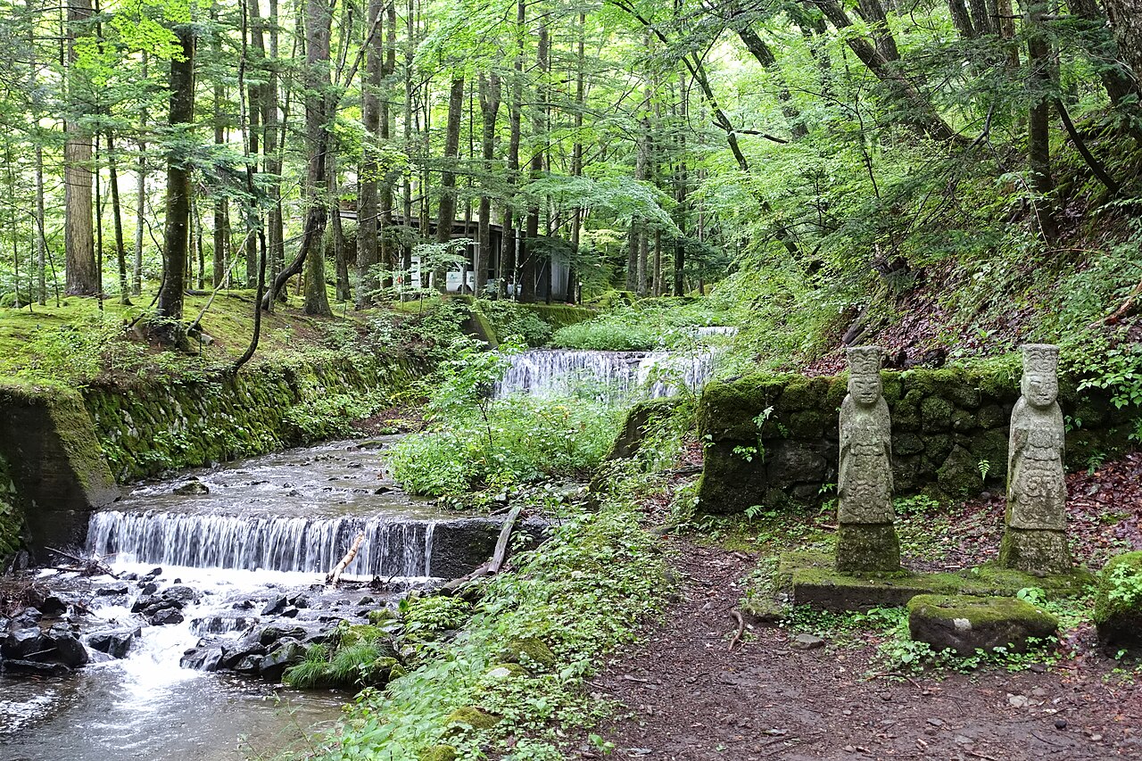 矢ヶ崎川の浅い流れと、川沿いの木立に並ぶ二体の石像