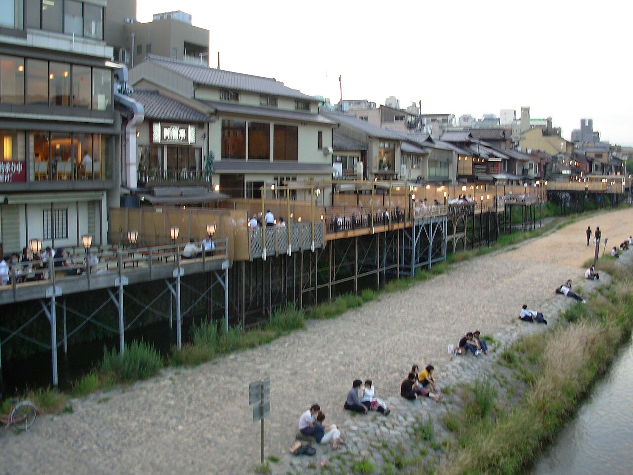 夕暮れの鴨川。灯りのついた納涼床と、河原に座って夕涼みする人々