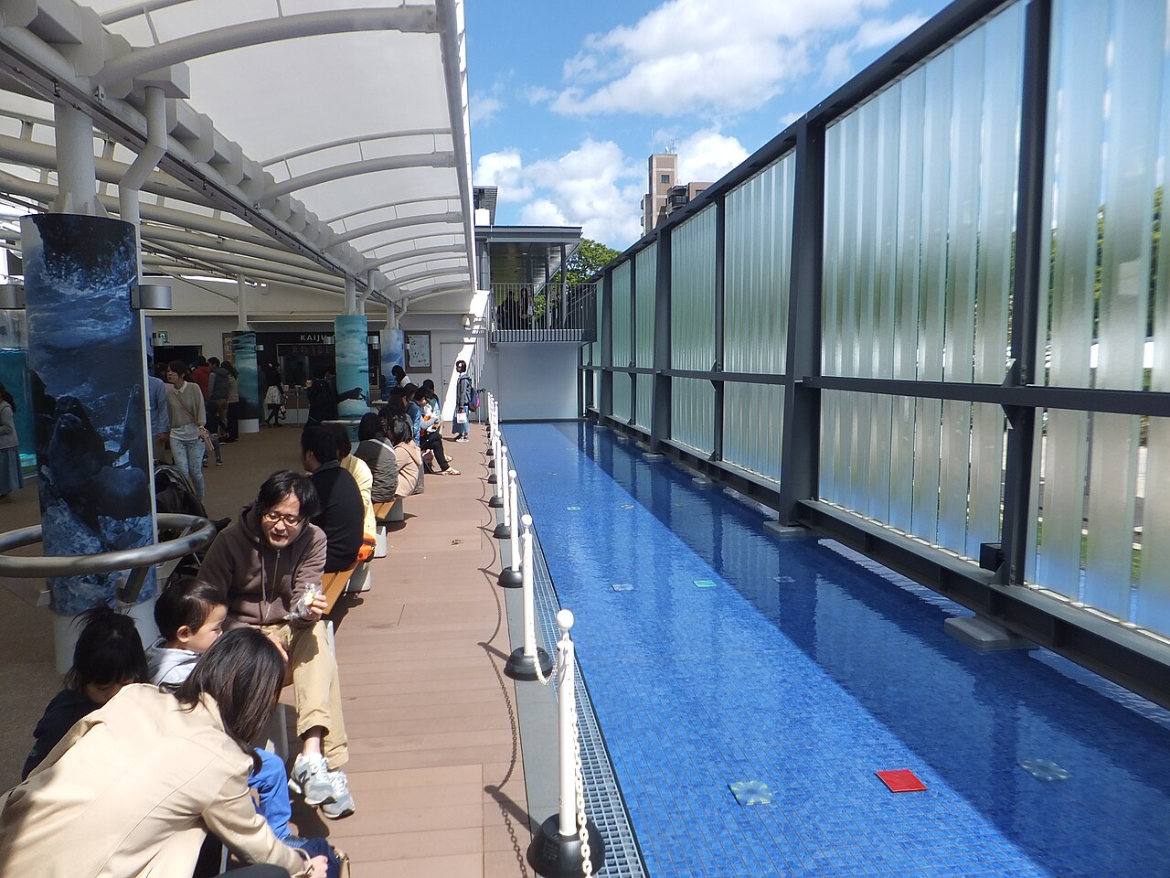 京都水族館の屋外通路と青い水路、日除けの屋根の下に座る来館者
